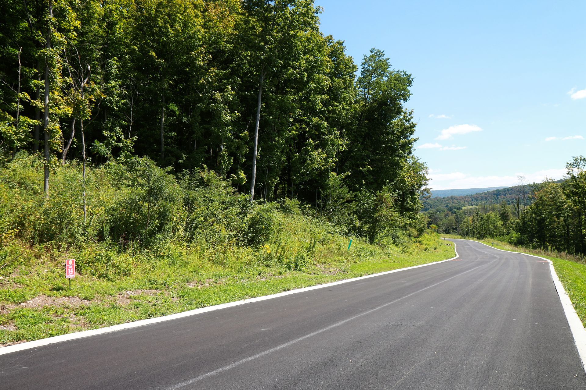 Paved road curving through a green, wooded landscape under a bright blue sky.
