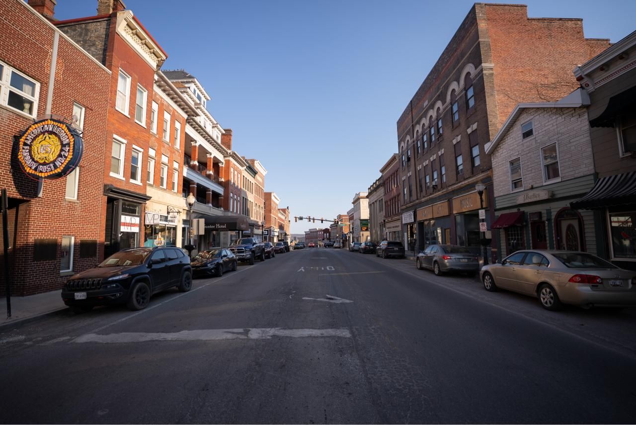 Street scene with brick buildings lining the road, cars parked along the sides, and a blue sky overhead.