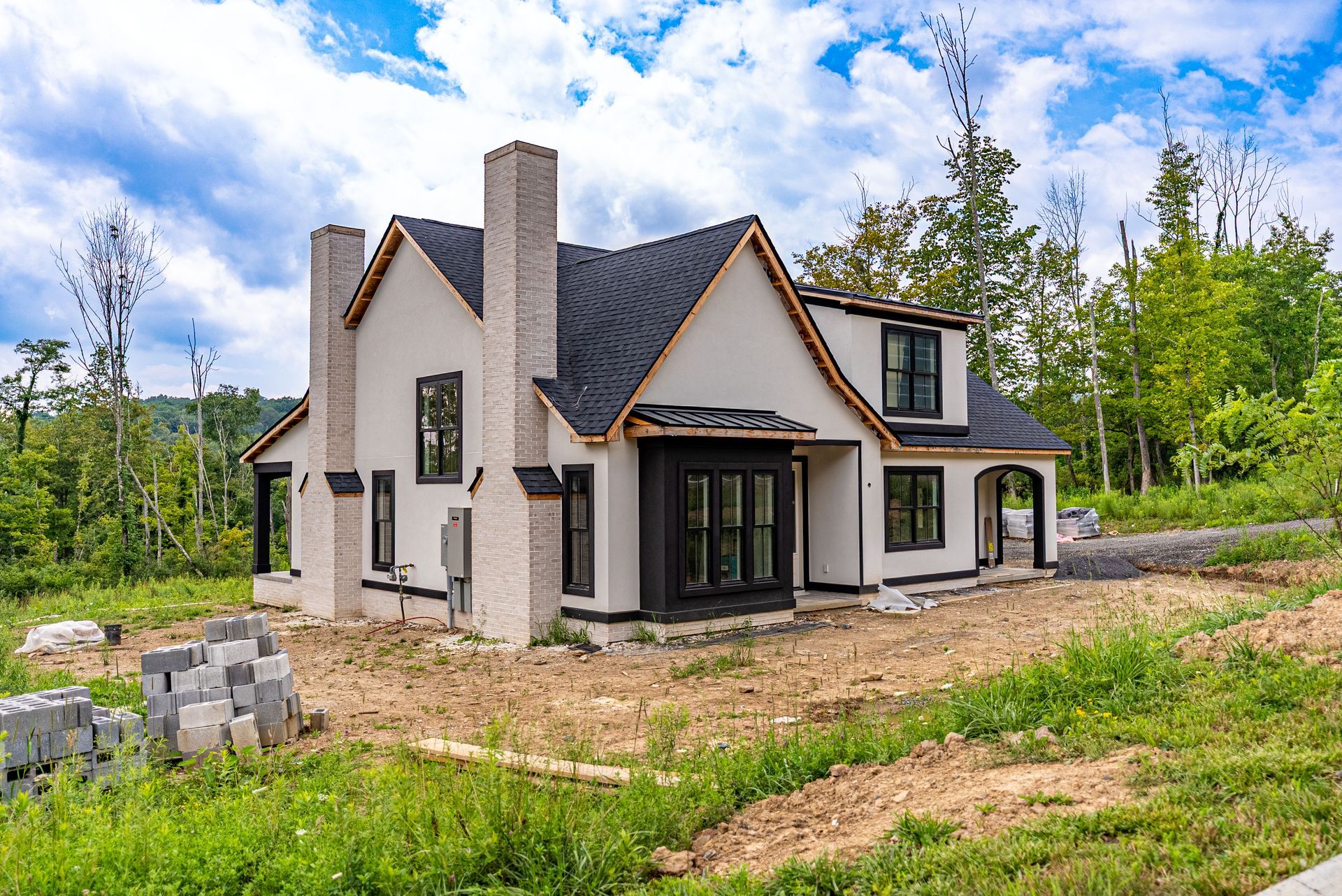 Newly constructed house with white stucco, black trim, and a black roof, set in a grassy, partially cleared lot under a cloudy sky.