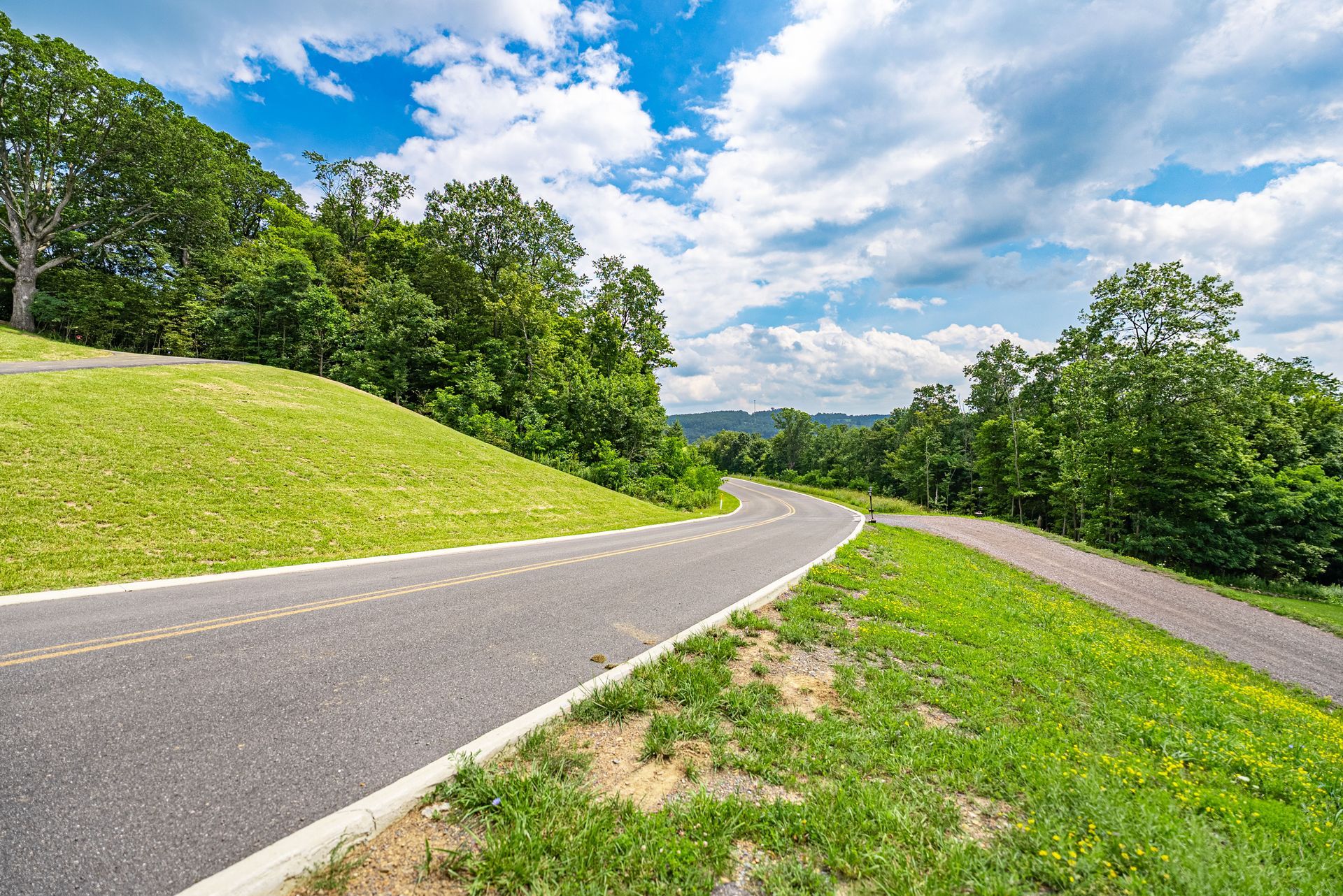 Paved road curves through a green landscape, flanked by grass and trees, under a blue sky with clouds.