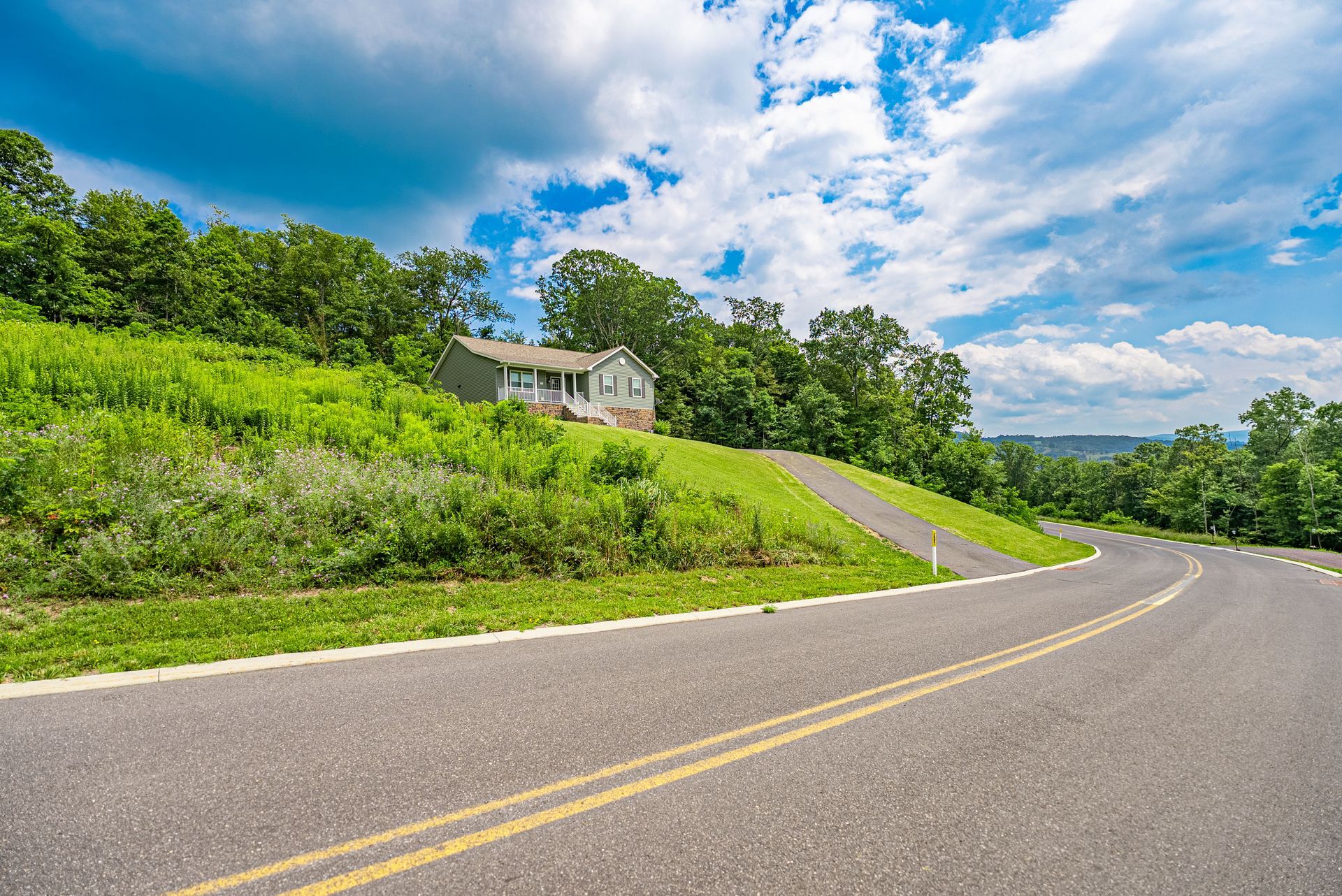 A small house sits on a grassy hillside next to a winding road under a partly cloudy blue sky.