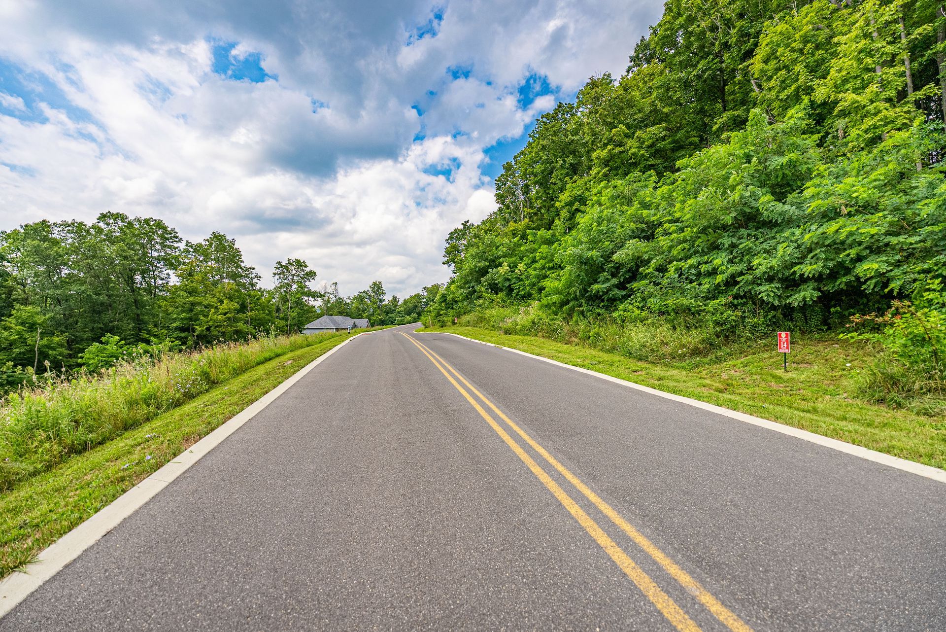 Paved road with double yellow lines winding through a lush green forest under a partly cloudy sky.
