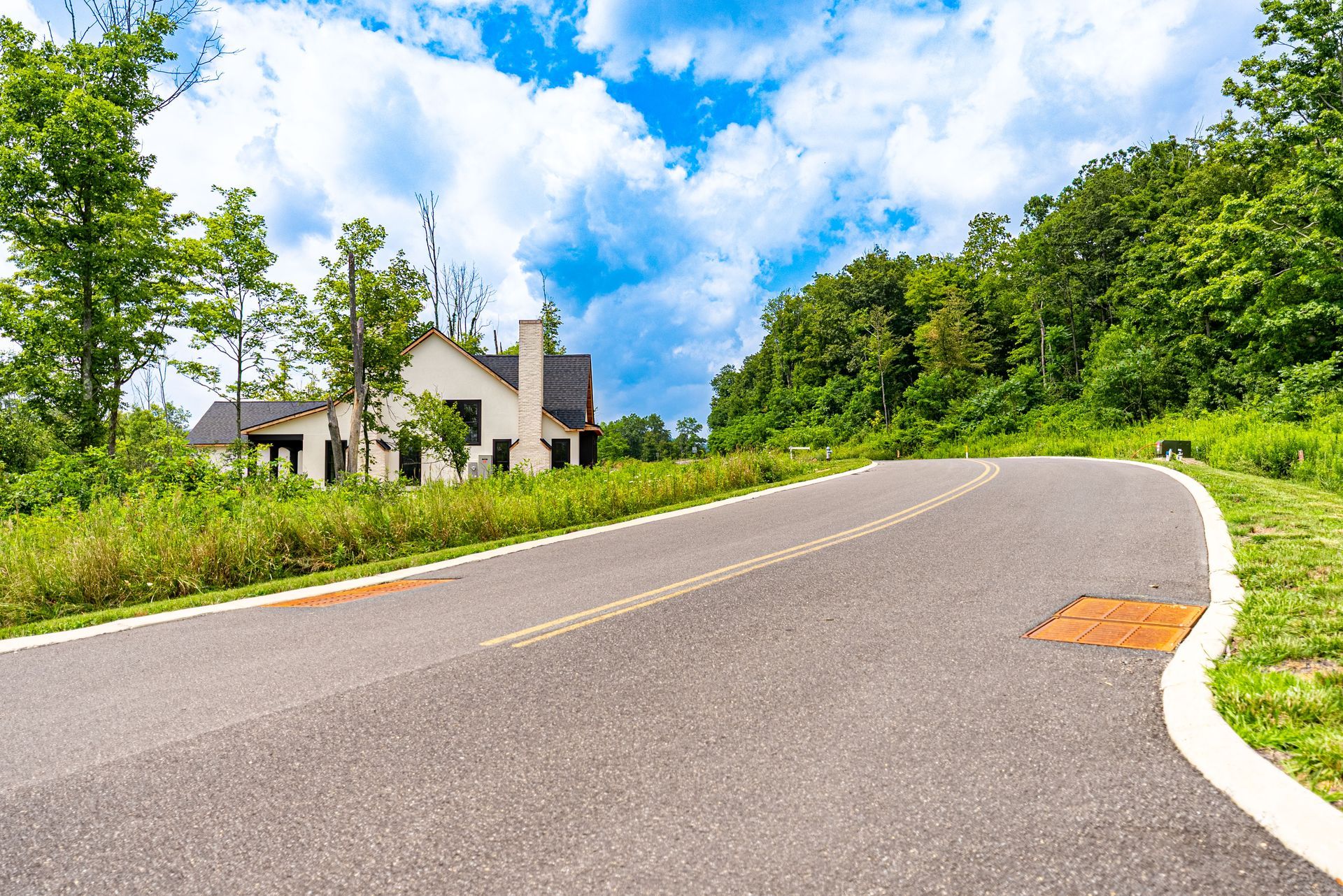 Paved road curves uphill toward a house under construction, bordered by grassy areas and a forested hillside on a bright, cloudy day.