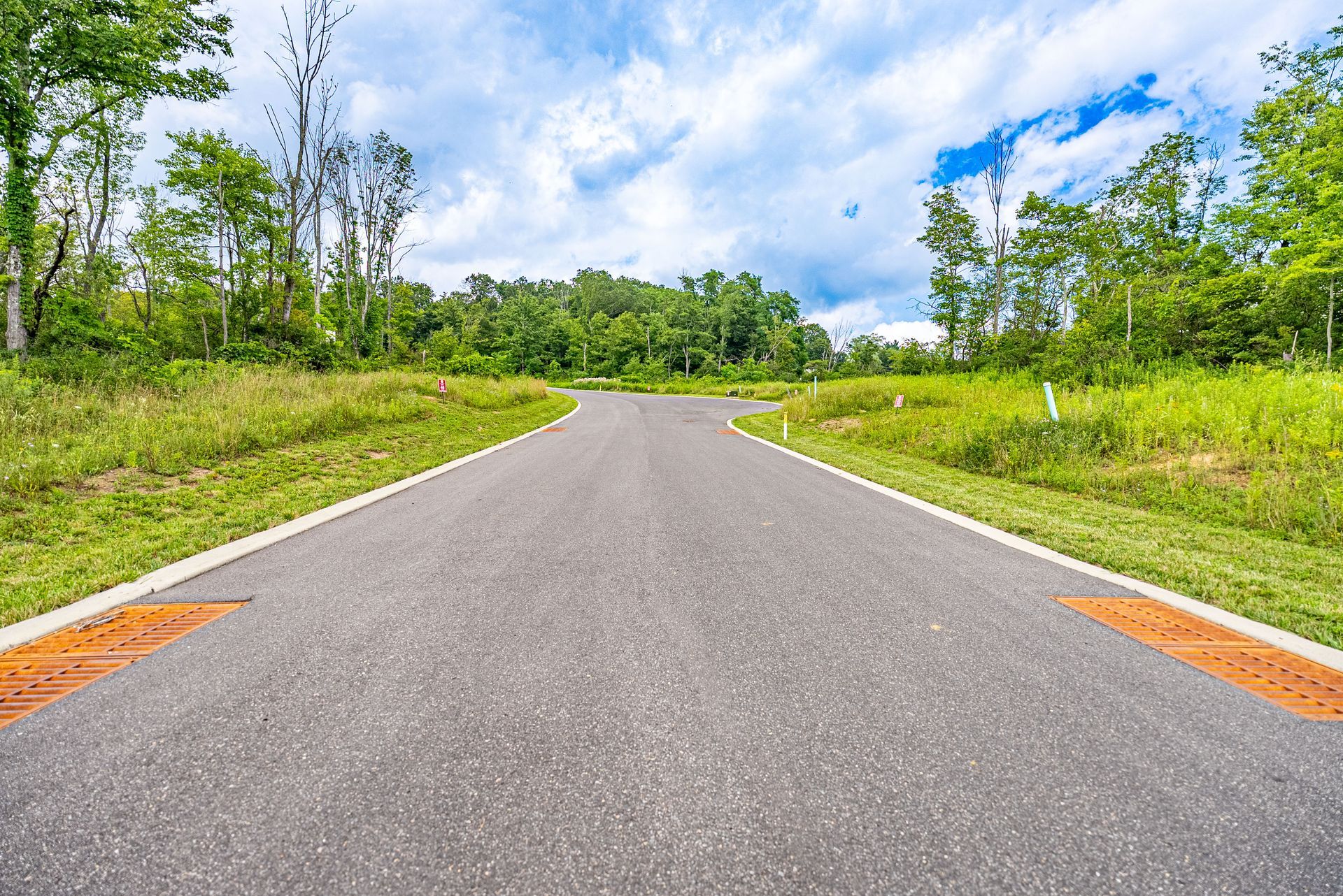 Paved road leading through grassy landscape towards a treeline under a partly cloudy sky.
