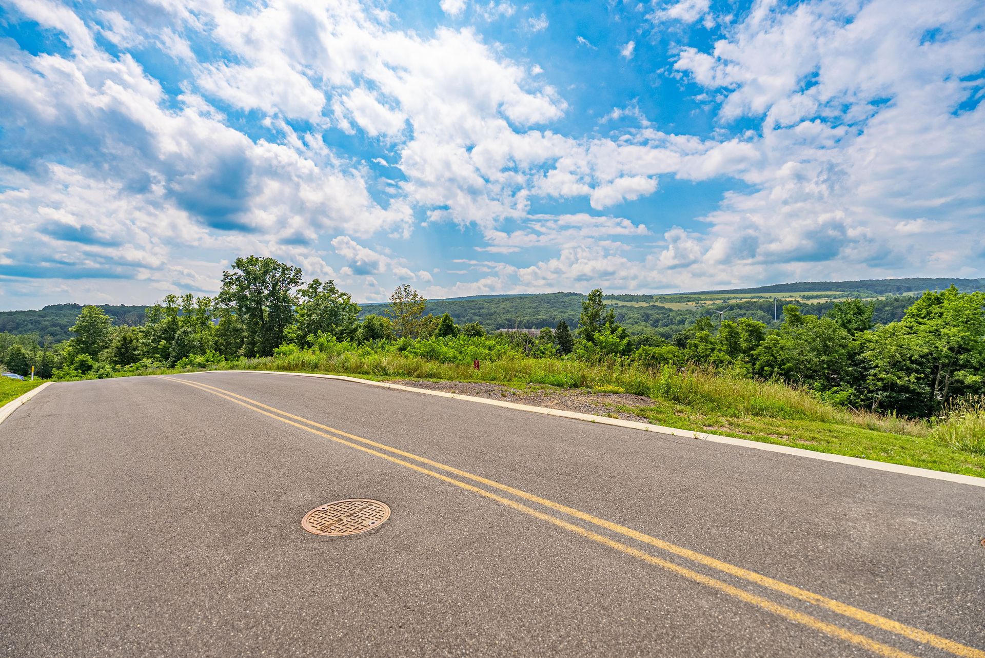 Asphalt road curving downhill with a view of green trees and a cloudy blue sky.