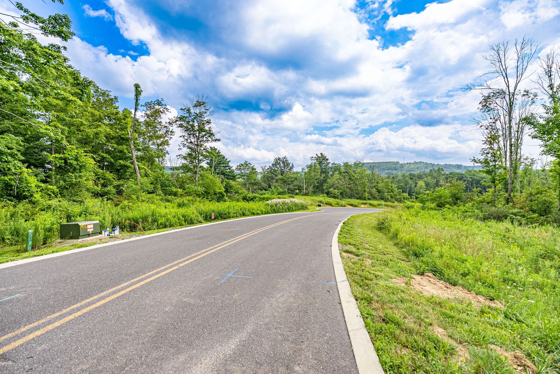 Winding asphalt road through a lush green landscape under a bright blue sky with scattered clouds.