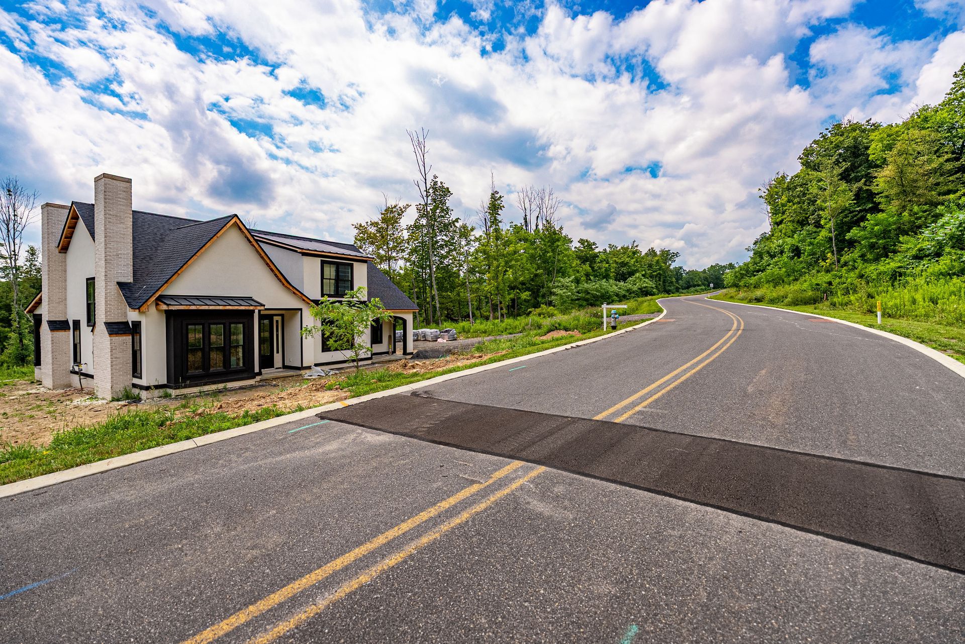 Modern house on a winding road, framed by lush greenery. The sky is blue with fluffy white clouds.