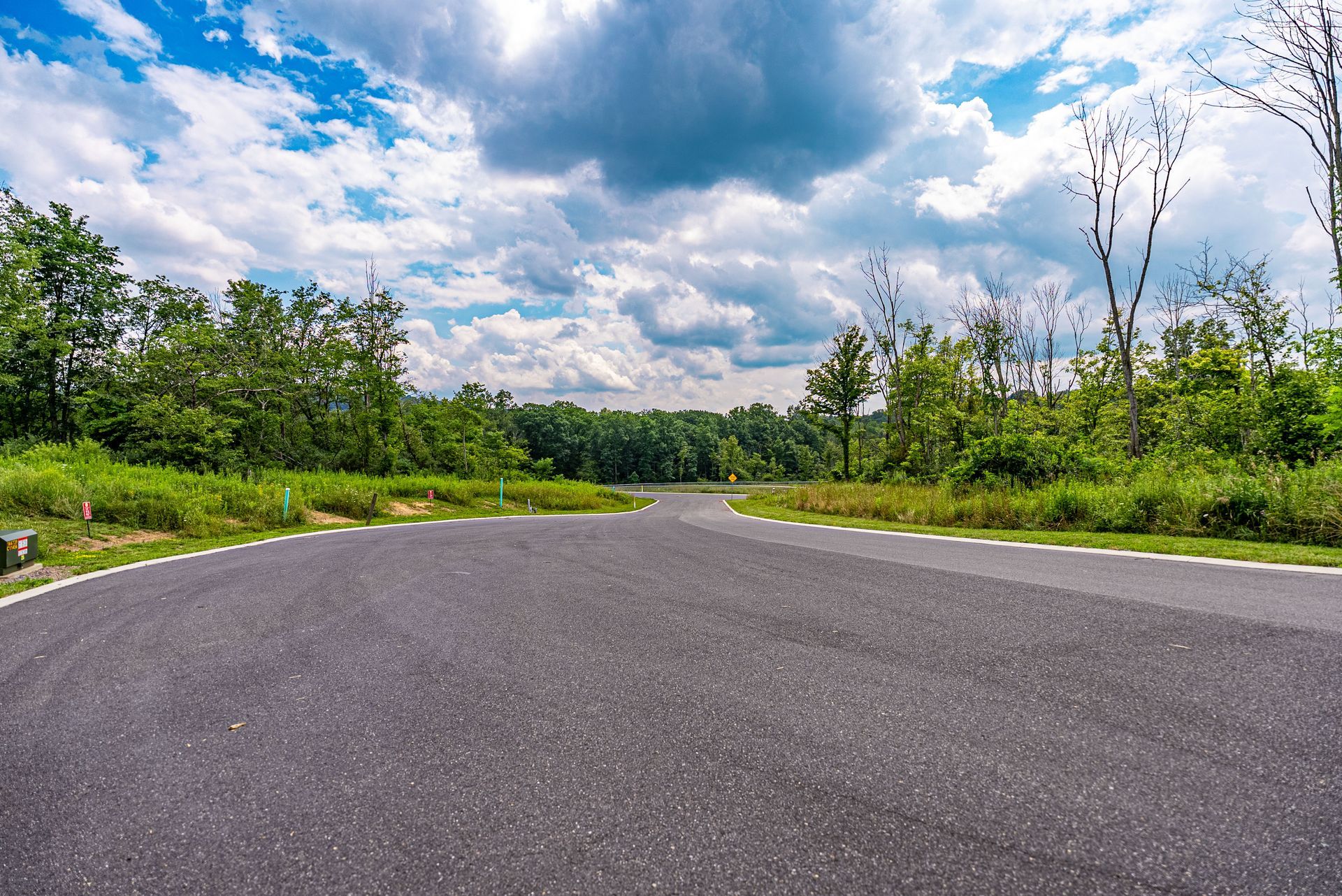 Paved road splitting, surrounded by green trees and grass, under a cloudy blue sky.