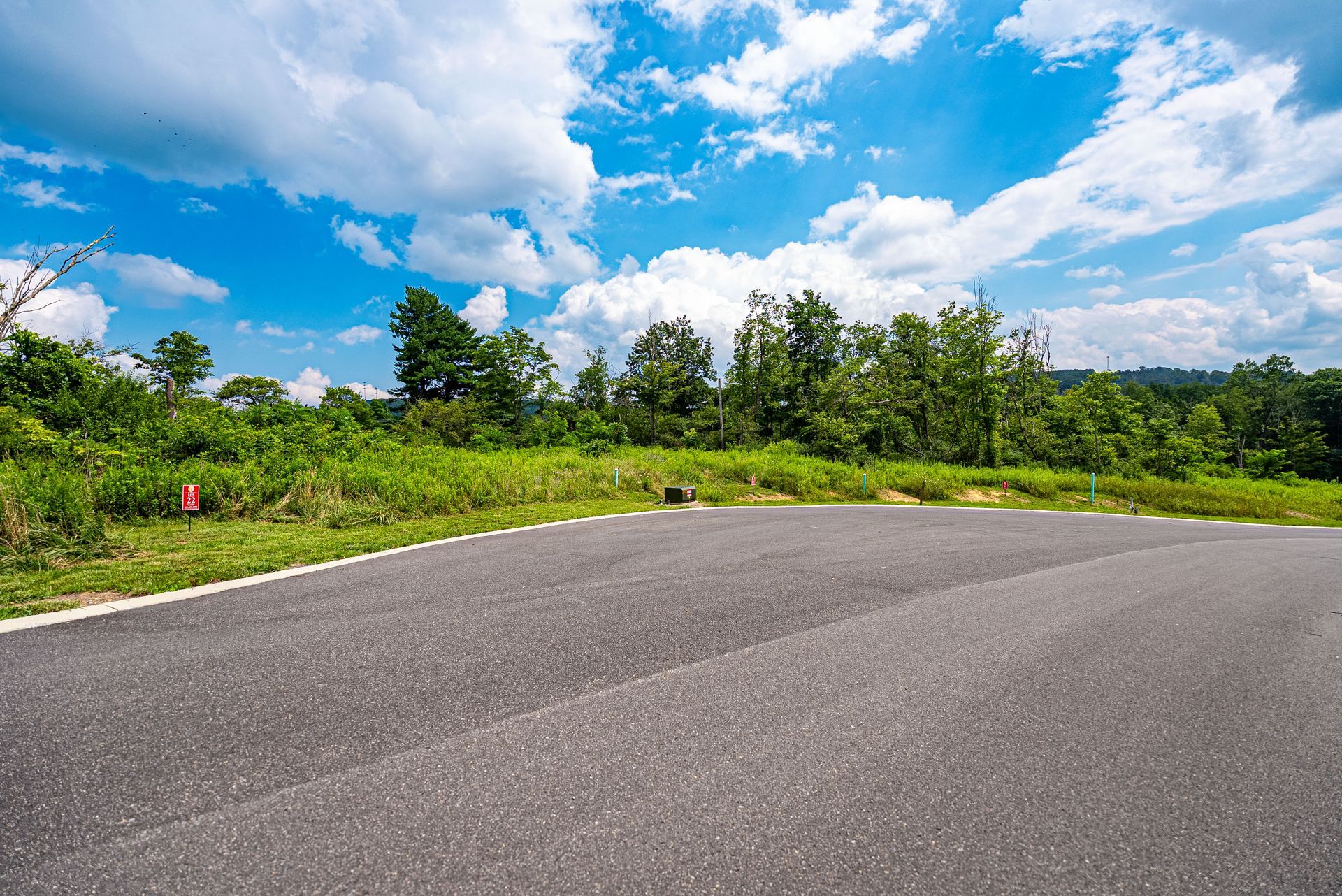 A paved road curves toward a green, grassy area with trees and a bright blue sky filled with white clouds.