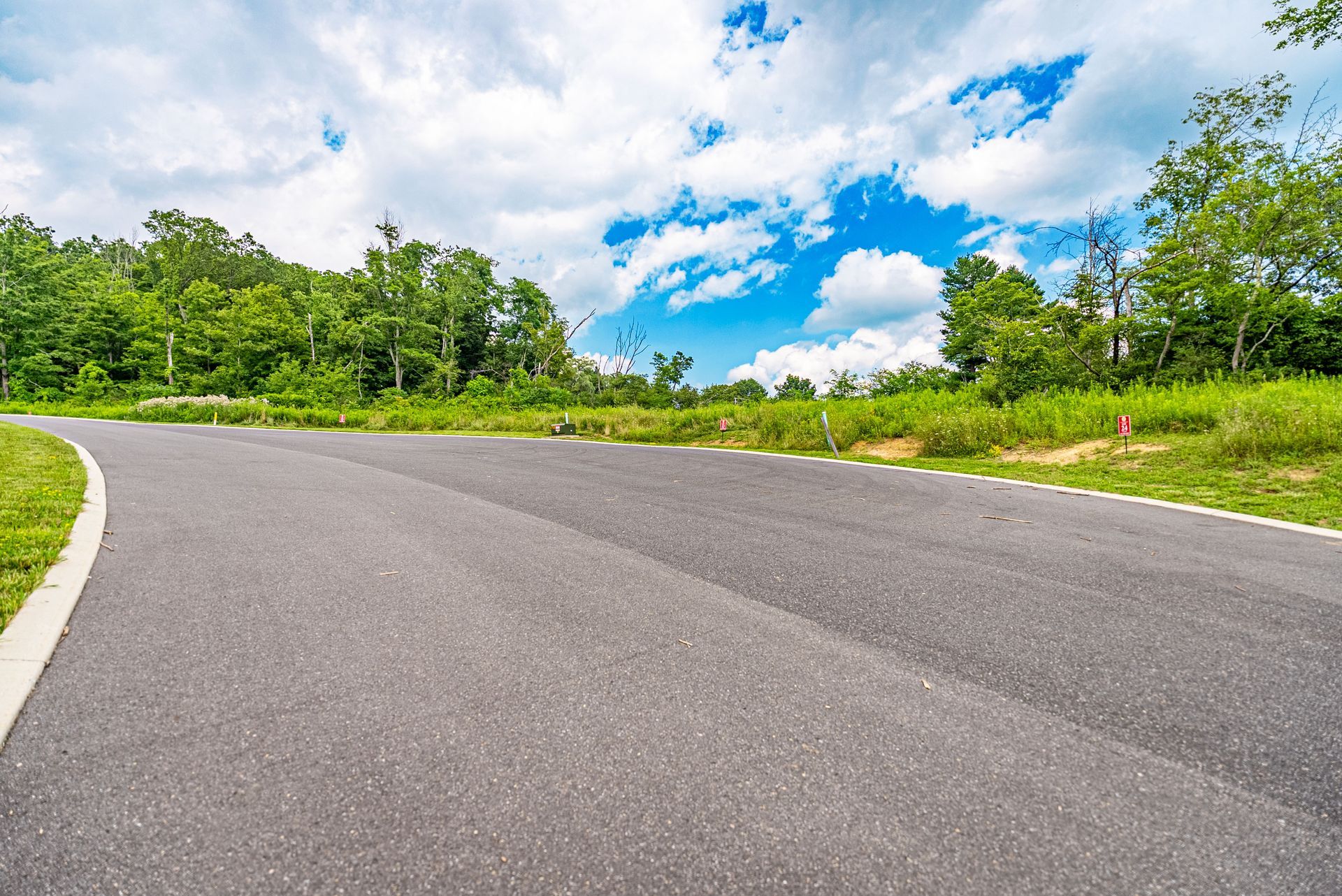 Curving asphalt road bordered by green grass and trees under a partly cloudy blue sky.