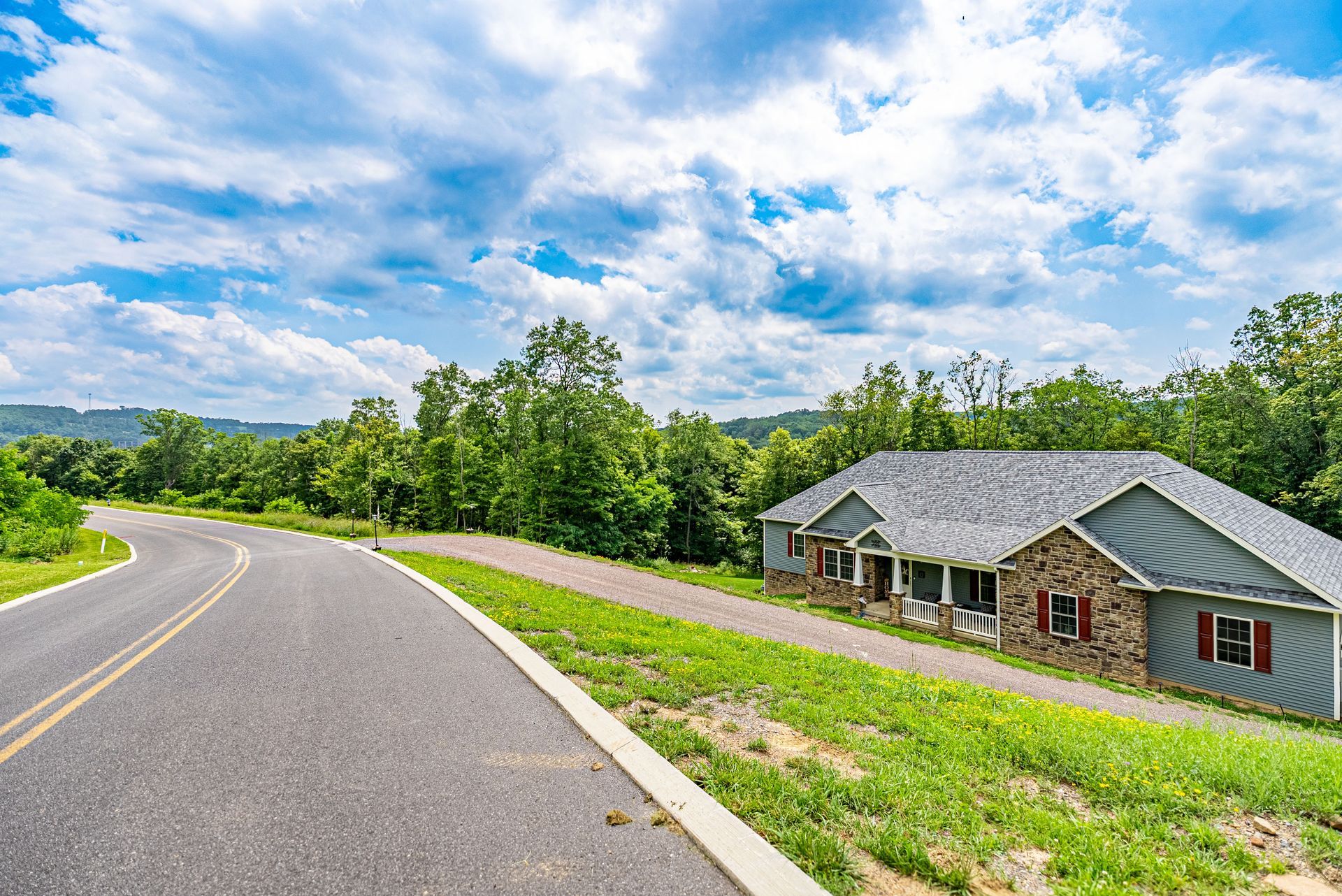 A house with a stone facade and gray siding sits beside a winding road. Green grass lines the road with trees and blue sky in the background.