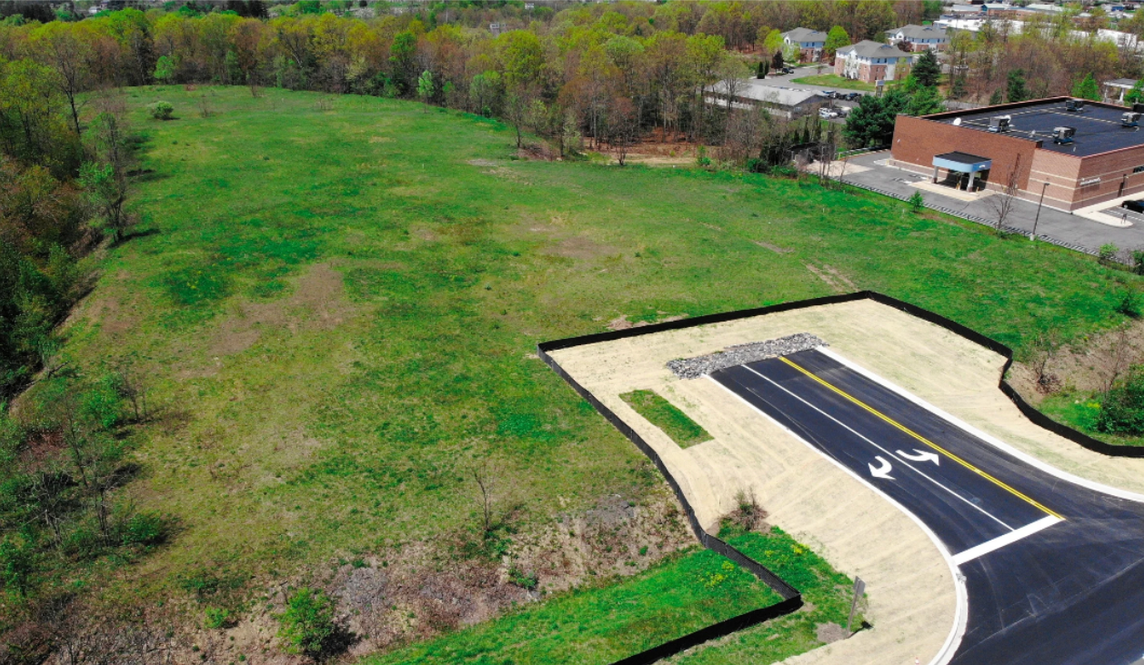 Aerial view of an empty grassy lot next to a newly paved road, bordered by trees and a building.