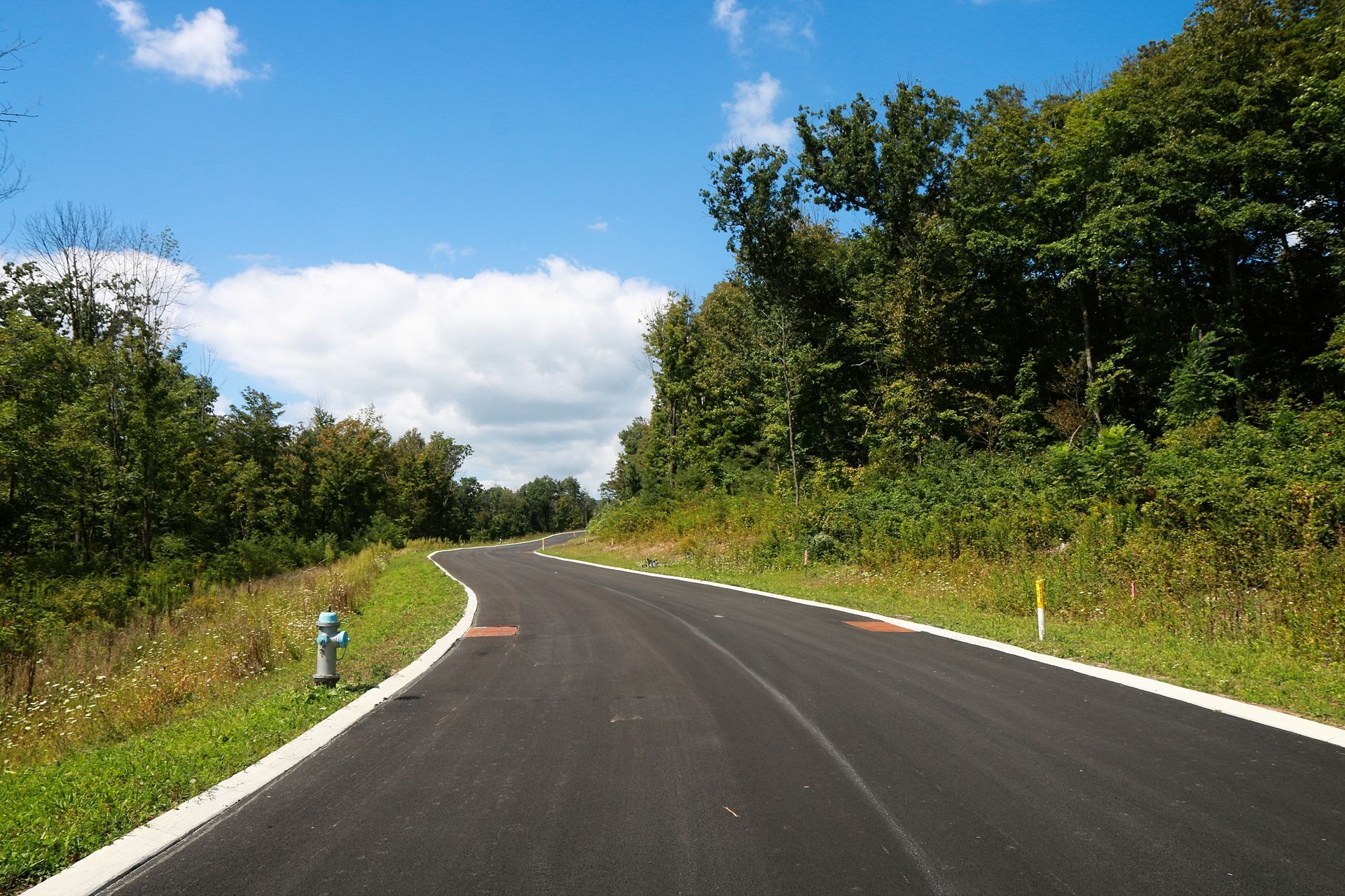 A winding asphalt road curves through a lush green forest under a partly cloudy blue sky.