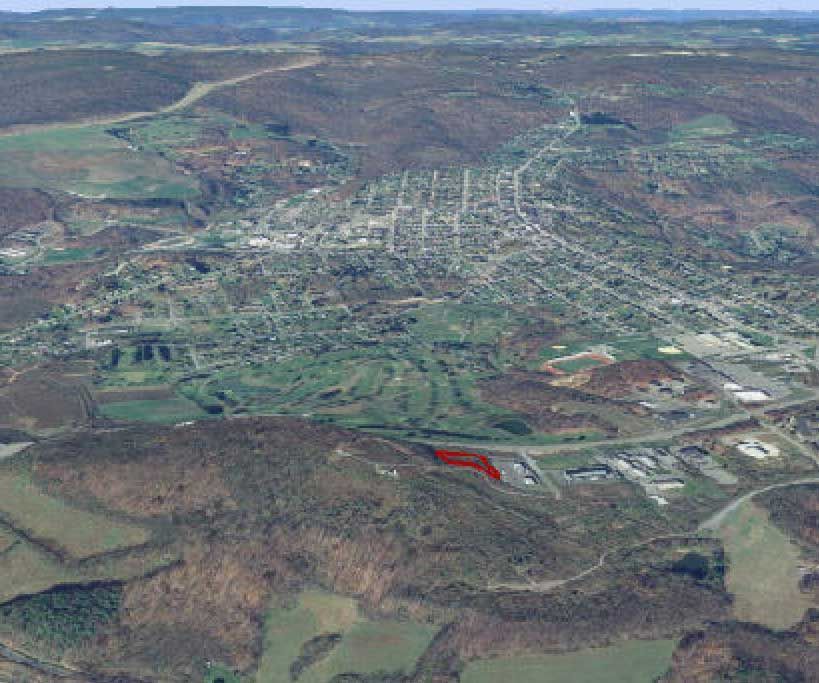 Aerial view of a town in a valley, with a red-highlighted area indicating a potential site; forested hills in the background.
