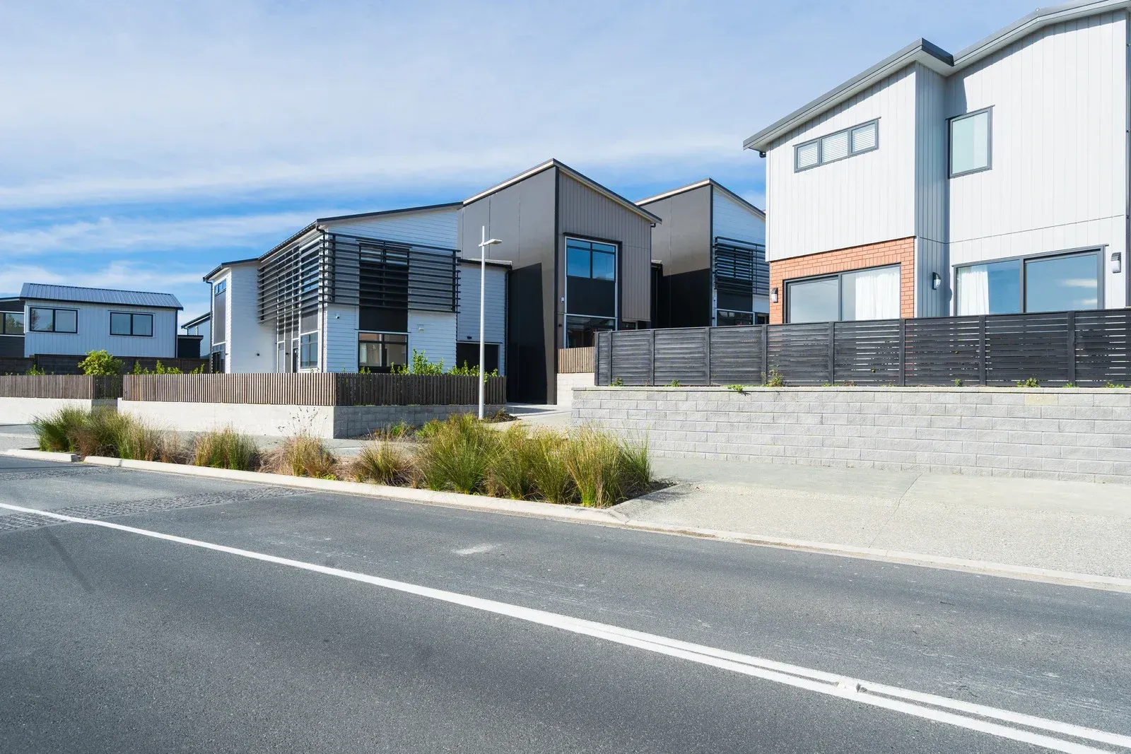 Modern houses with gray and wood siding, along a street with a median of bushes. Blue sky.