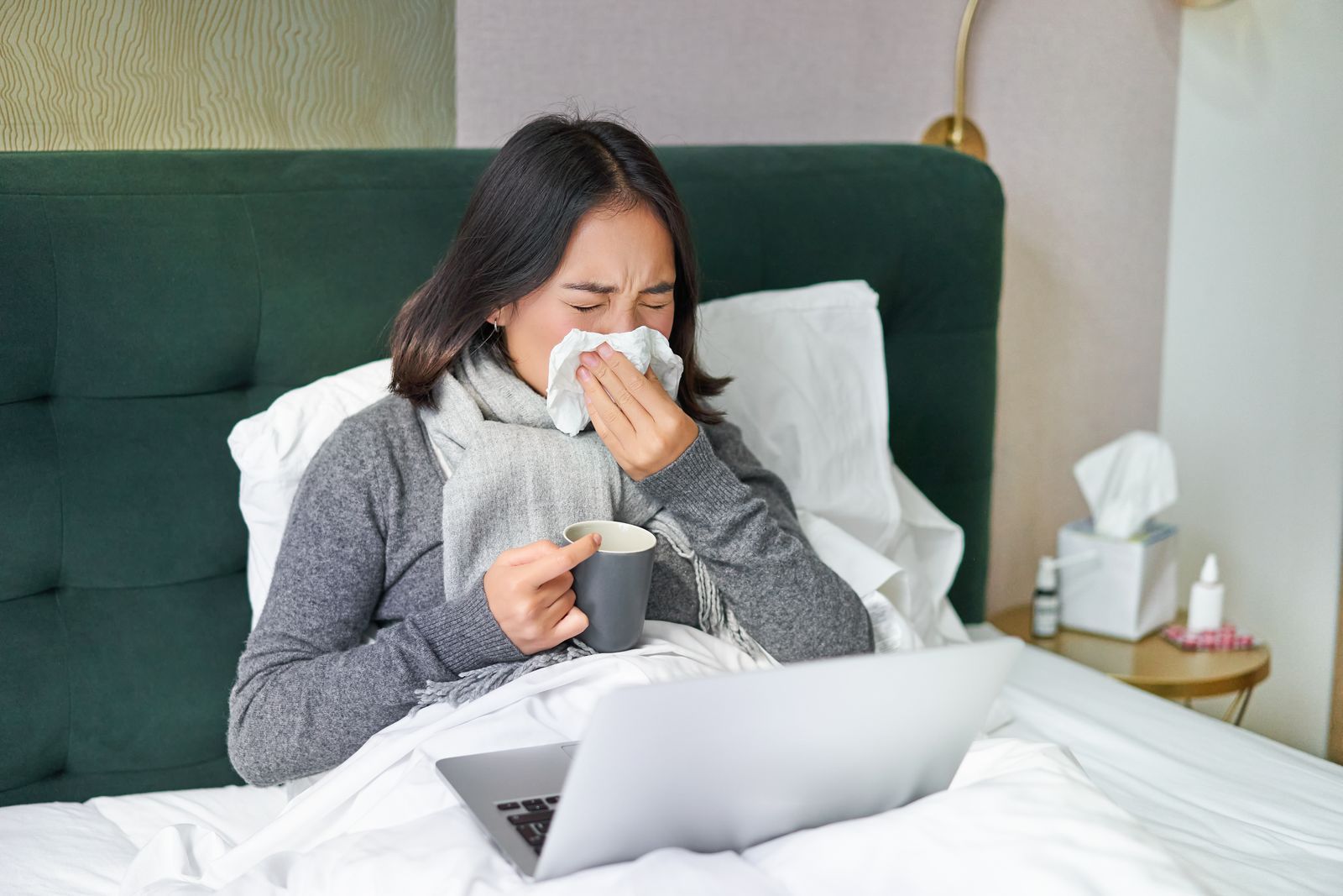 A woman is laying in bed with a laptop and blowing her nose.