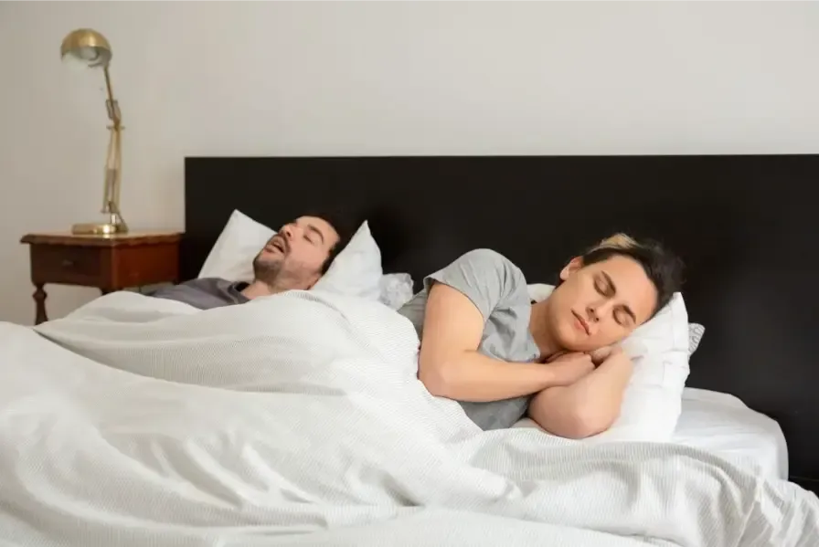 A couple sleeping in bed, with one person snoring while the other struggles to sleep