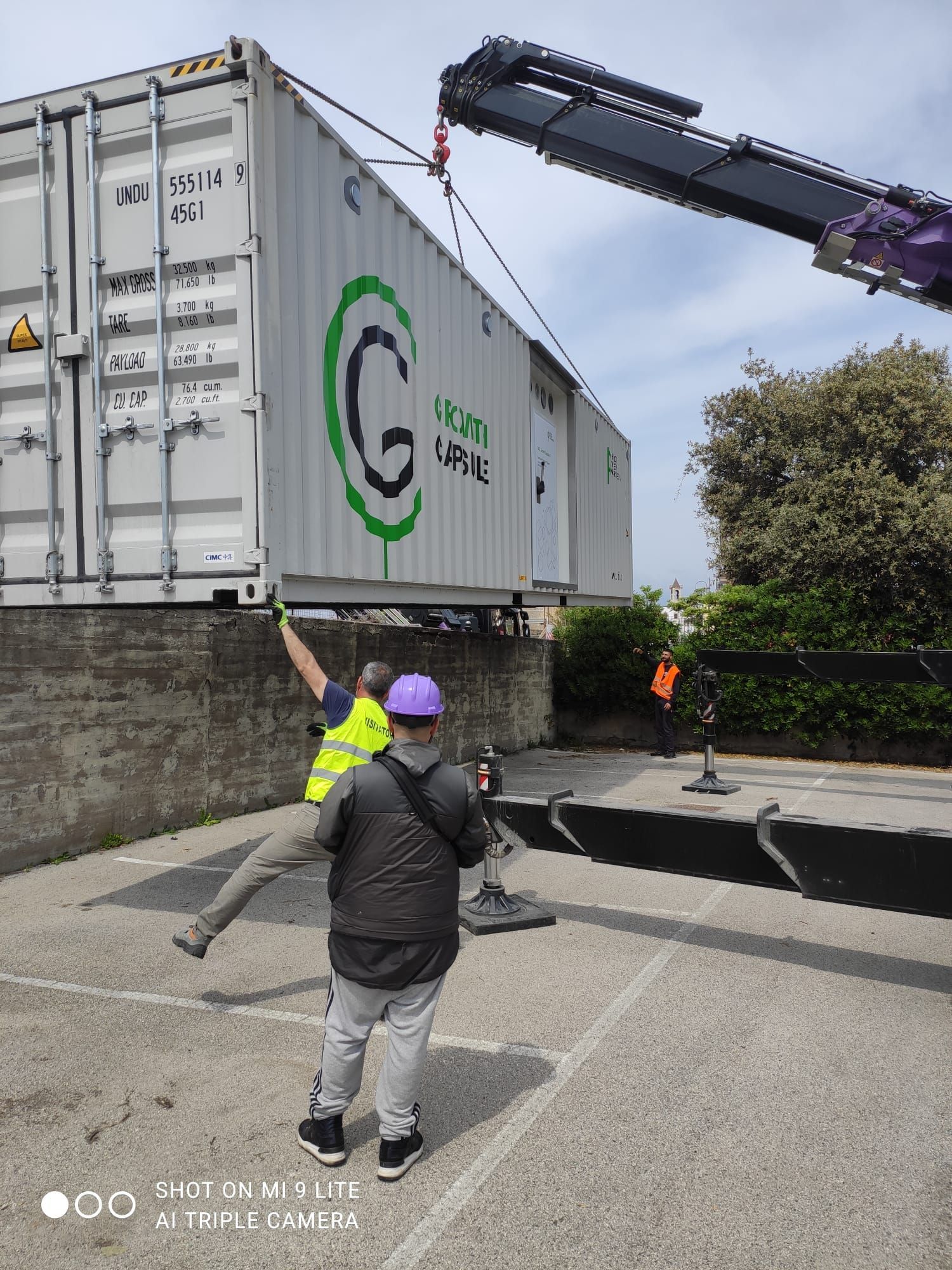 Una gru solleva un container con l'assistenza di due operai. Il container ha un logo verde, all'esterno.