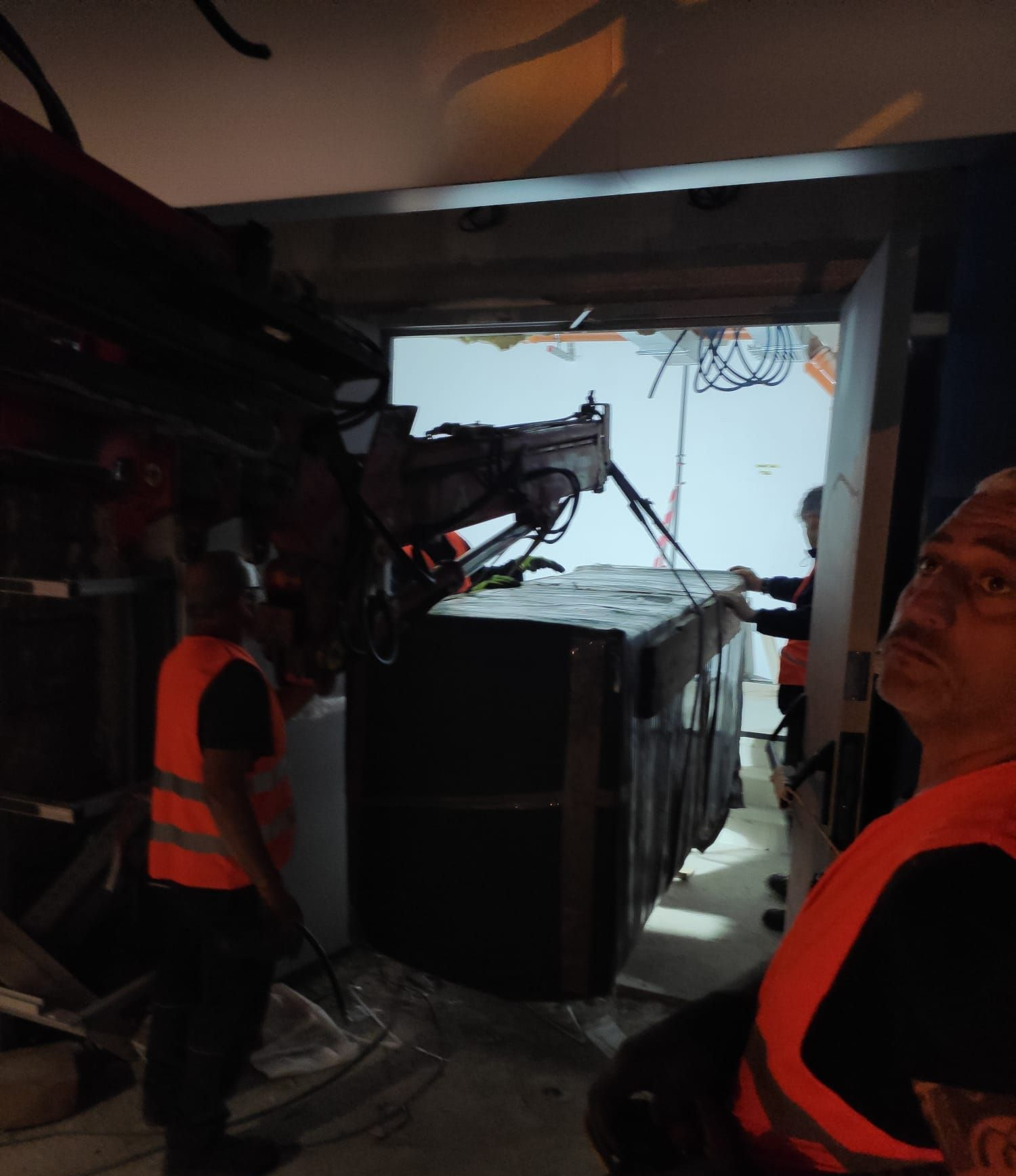 Gli operai con i giubbotti di sicurezza utilizzano una gru per sollevare una grande cassa avvolta e farla entrare in una porta.