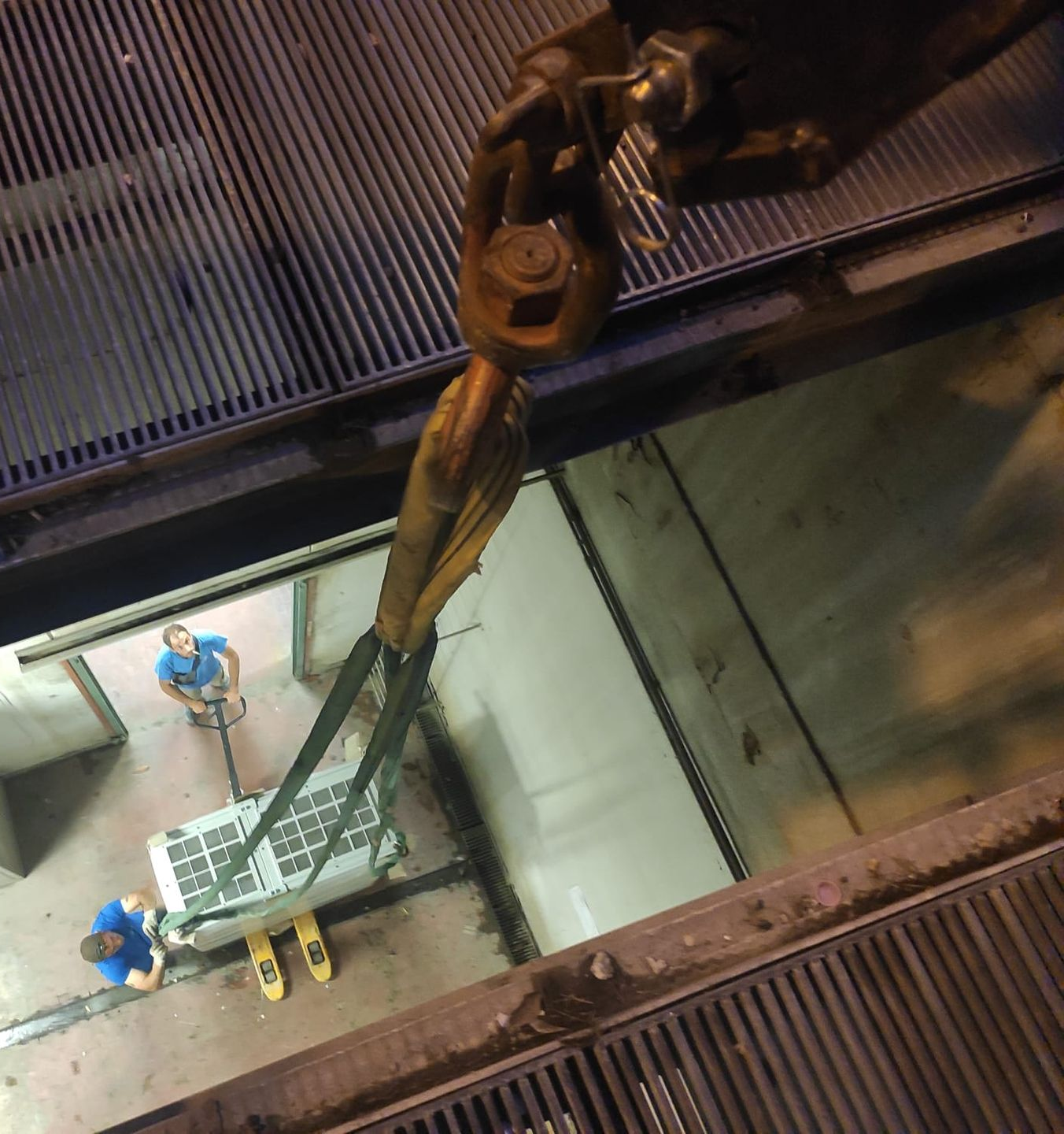 Gru che solleva una cassa rettangolare tra due operai all'interno di una grande struttura industriale.