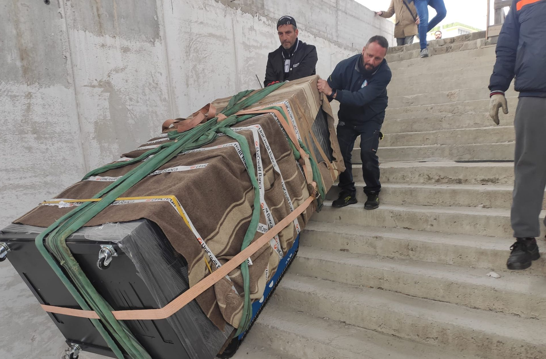 Degli uomini spostano con cautela un grande oggetto avvolto su per le scale di cemento.