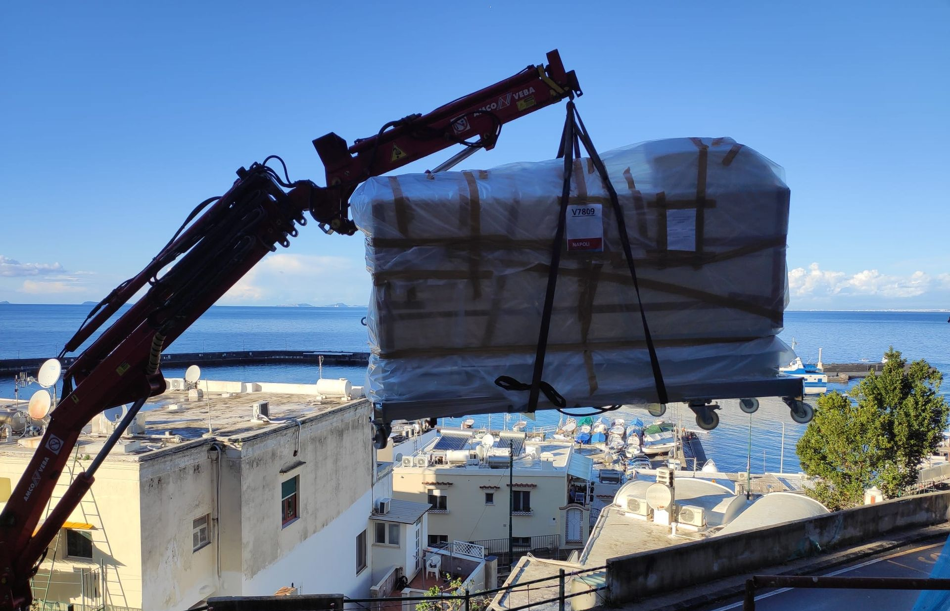 Una gru solleva un grande oggetto avvolto sopra una città costiera. Sullo sfondo, cielo azzurro e mare.