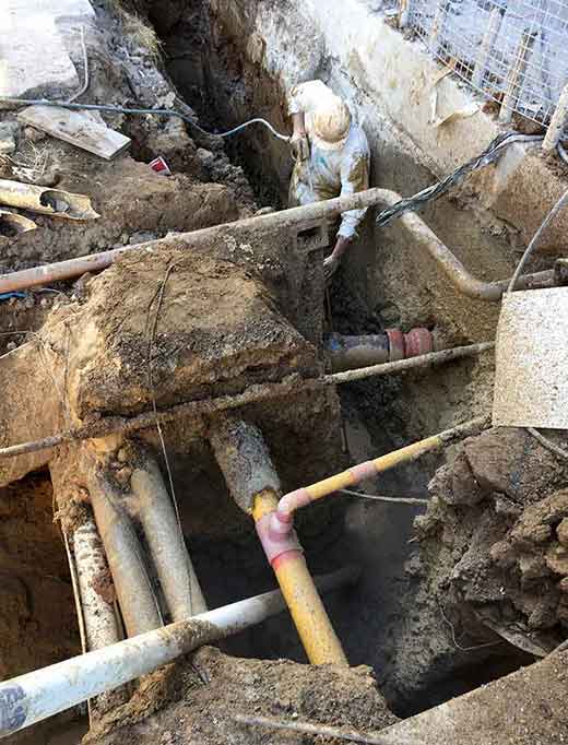 Worker in protective suit repairs pipes in an open trench; dirt, concrete, and various pipes are visible.