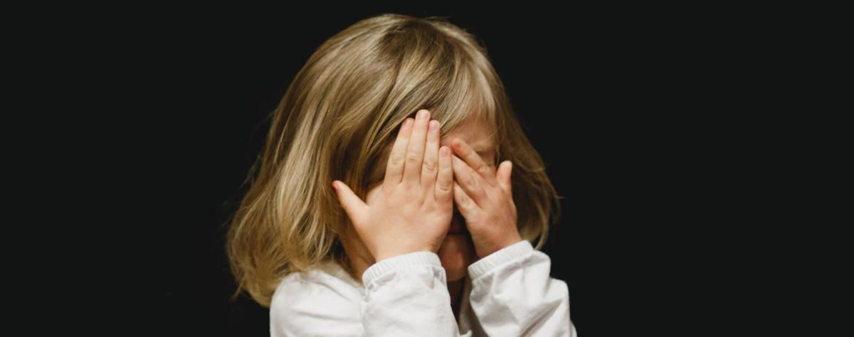 A little girl is covering her face with her hands on a black background.