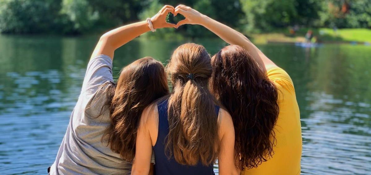 Three women are making a heart shape with their hands in front of a lake.