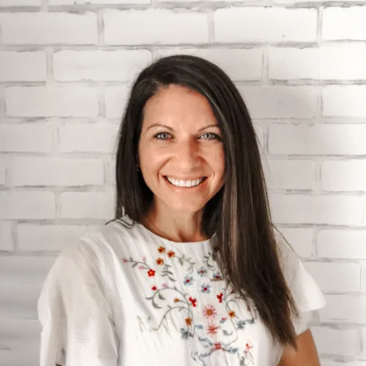 A woman in a white shirt is smiling in front of a white brick wall.