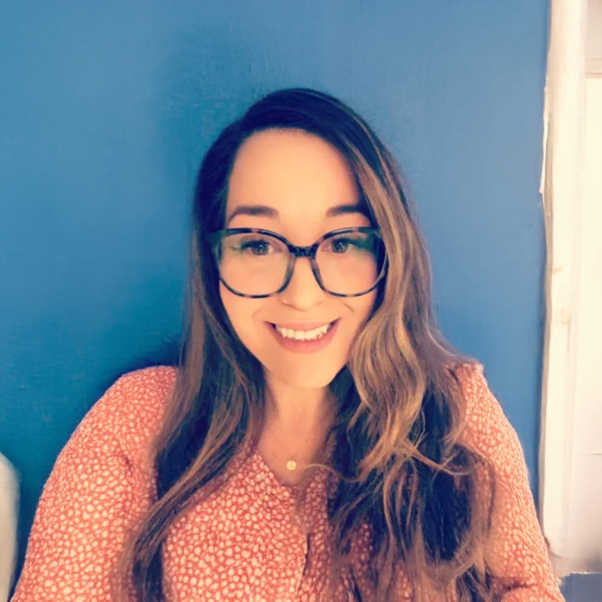 A woman wearing glasses and a pink shirt is smiling in front of a blue wall.