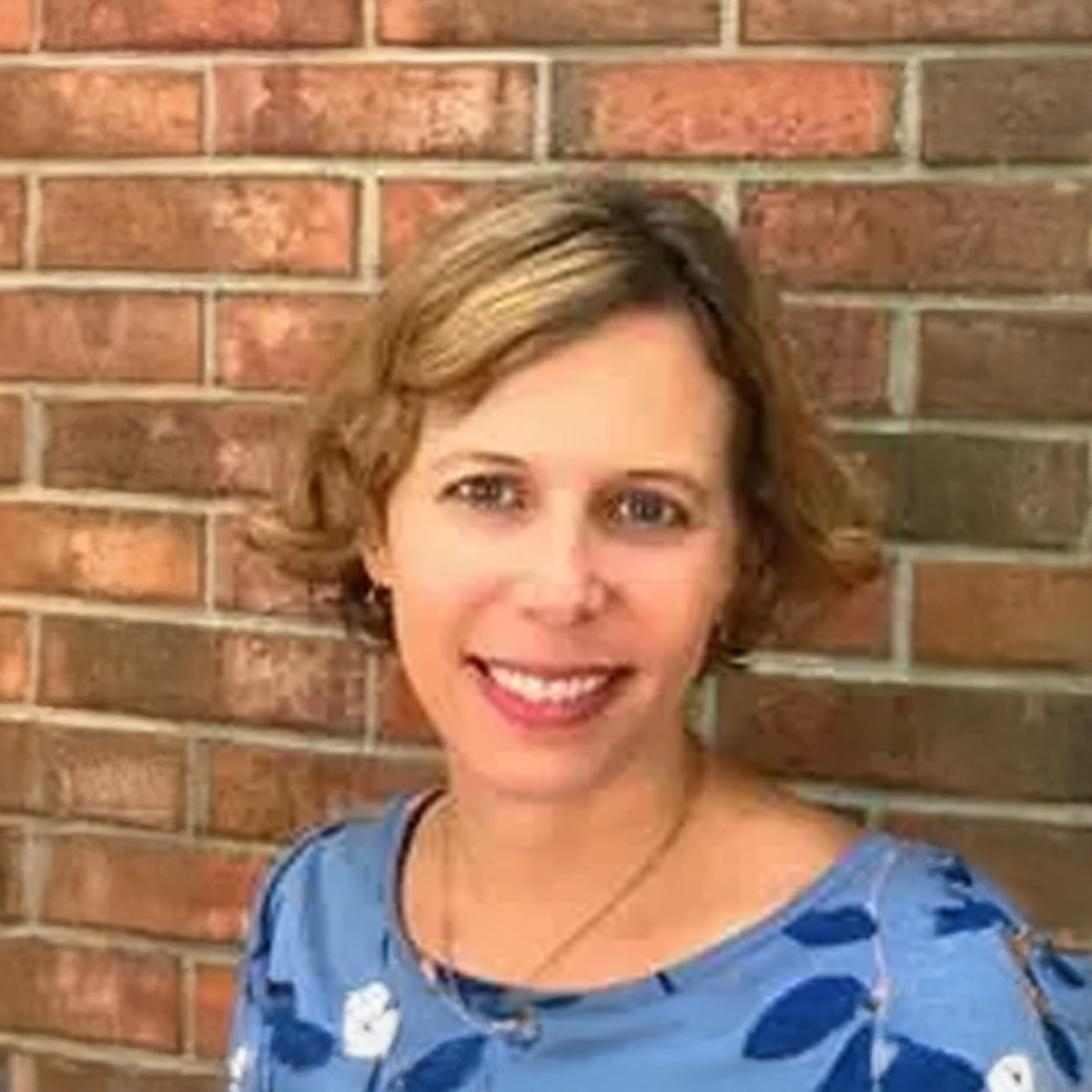 A woman in a blue shirt is smiling in front of a brick wall.