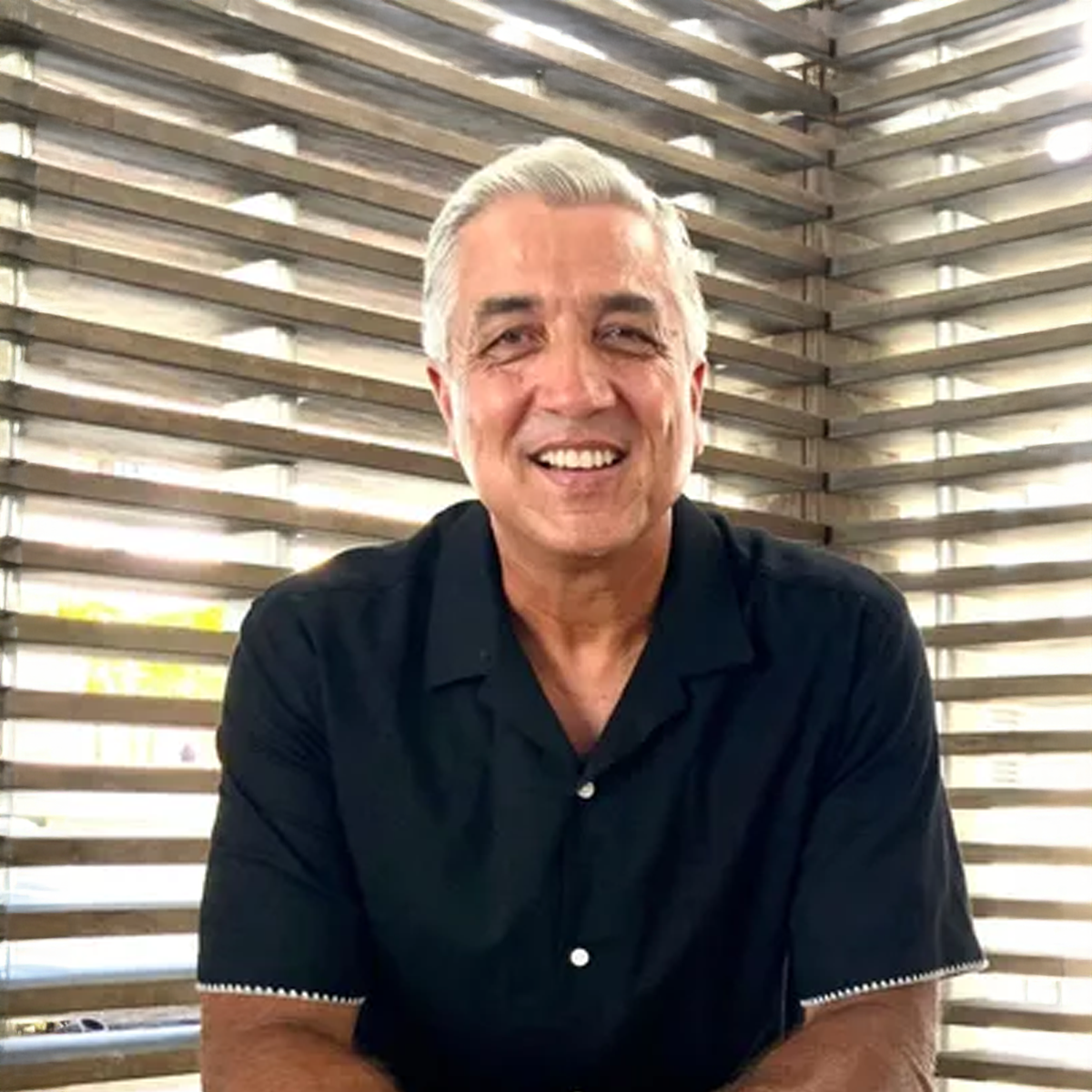 A man in a black shirt is sitting in front of a window and smiling.