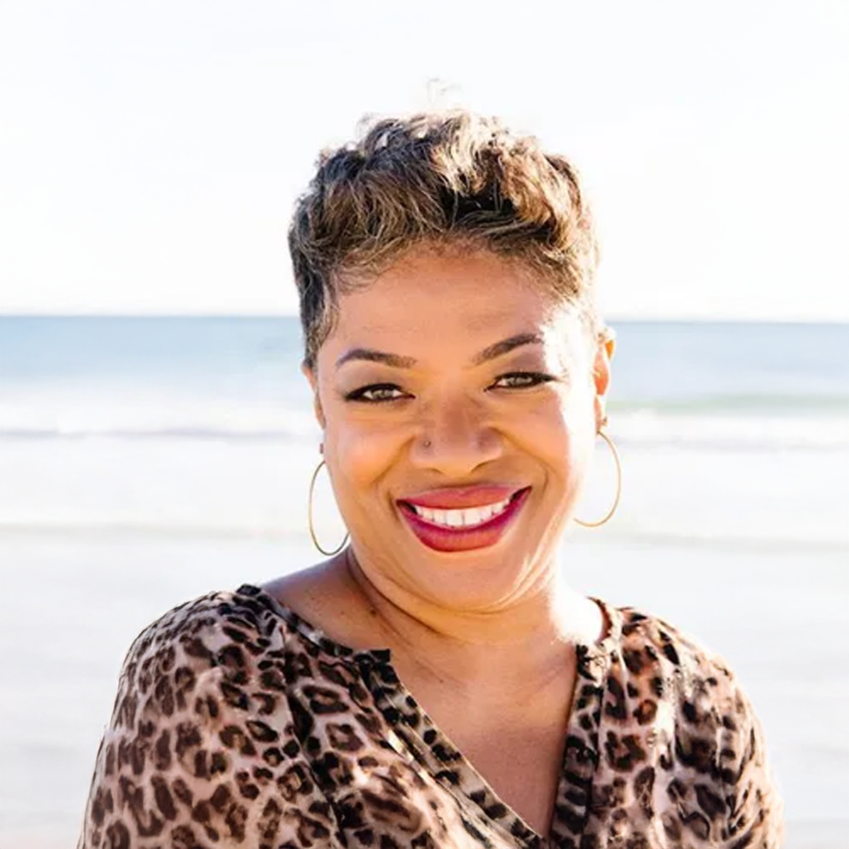 A woman in a leopard print shirt is smiling on the beach.