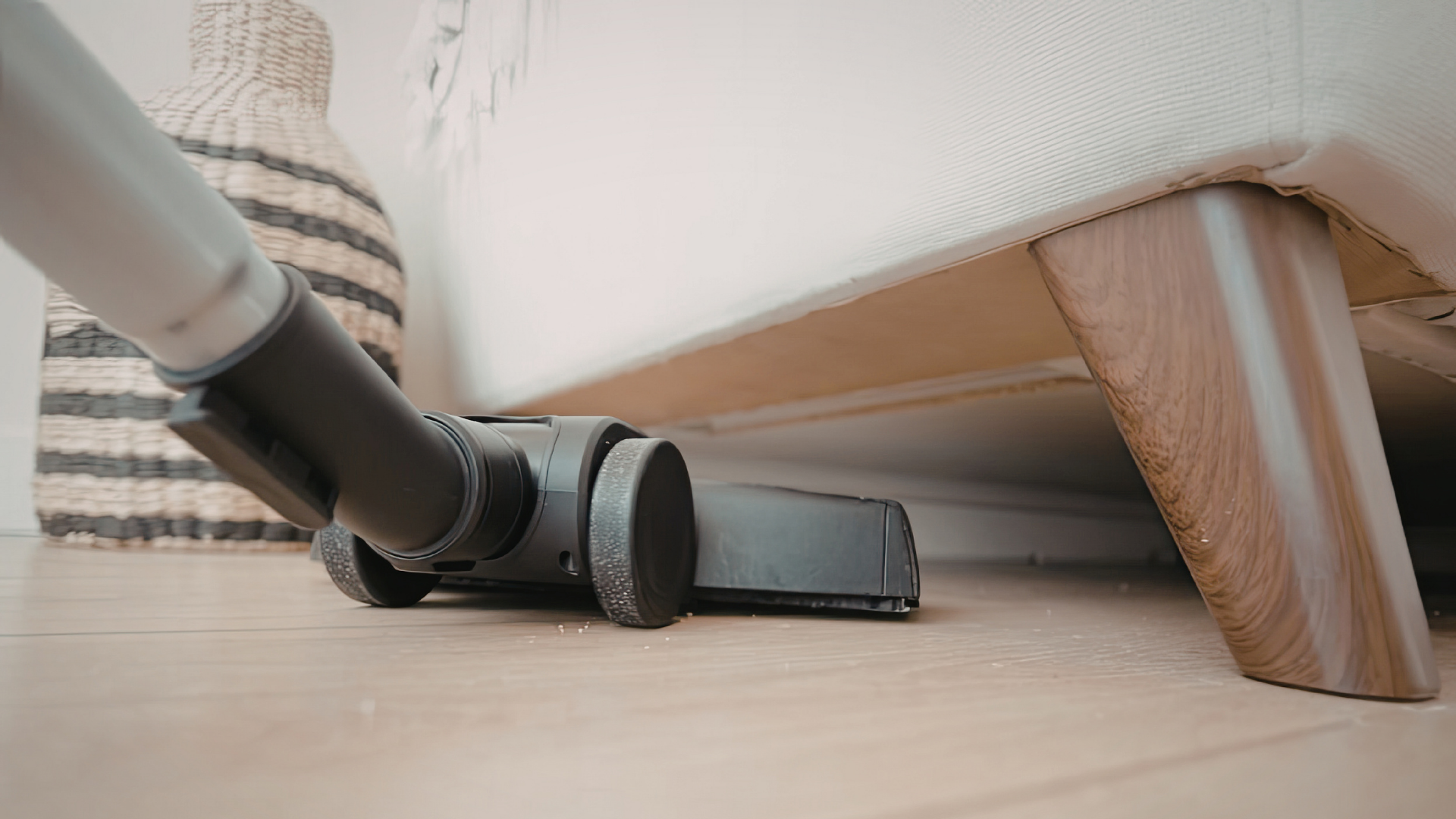 A vacuum cleaner attachment cleaning the floor beneath a light-colored sofa near a patterned basket.