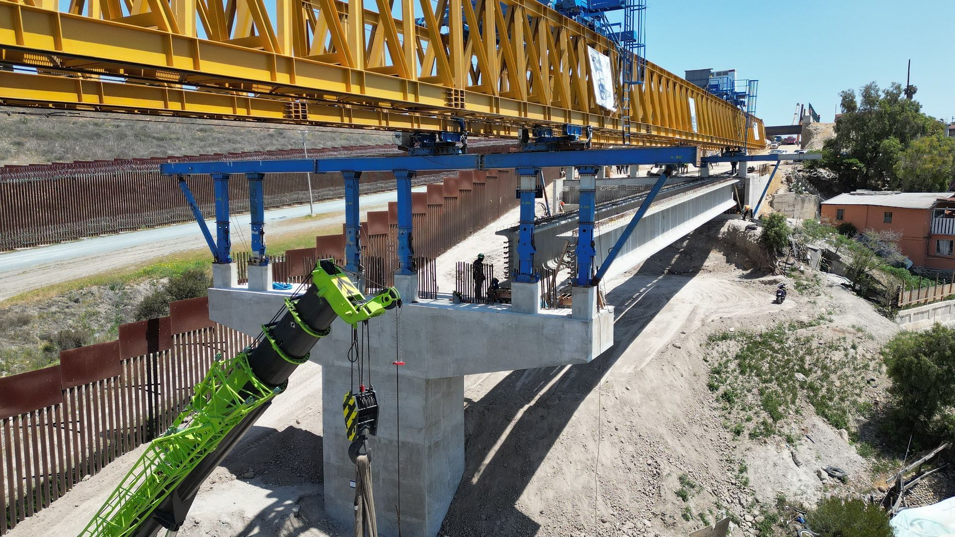 Construction of a concrete bridge. A crane lifts materials. Blue and yellow machinery. Outdoors.