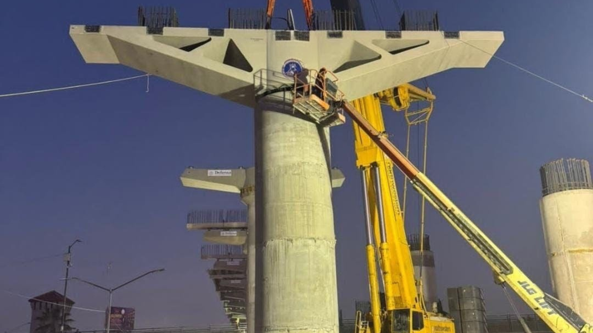 Construction site: Concrete bridge pier with workers in a lift, under a blue sky.