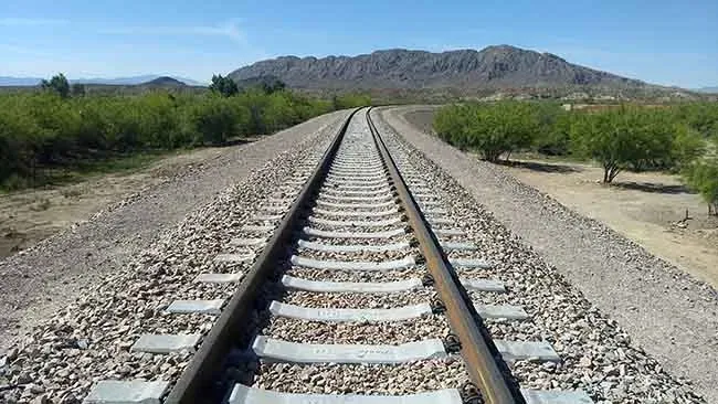 Railroad tracks leading toward a distant mountain under a clear blue sky; gravel bed, green trees on either side.
