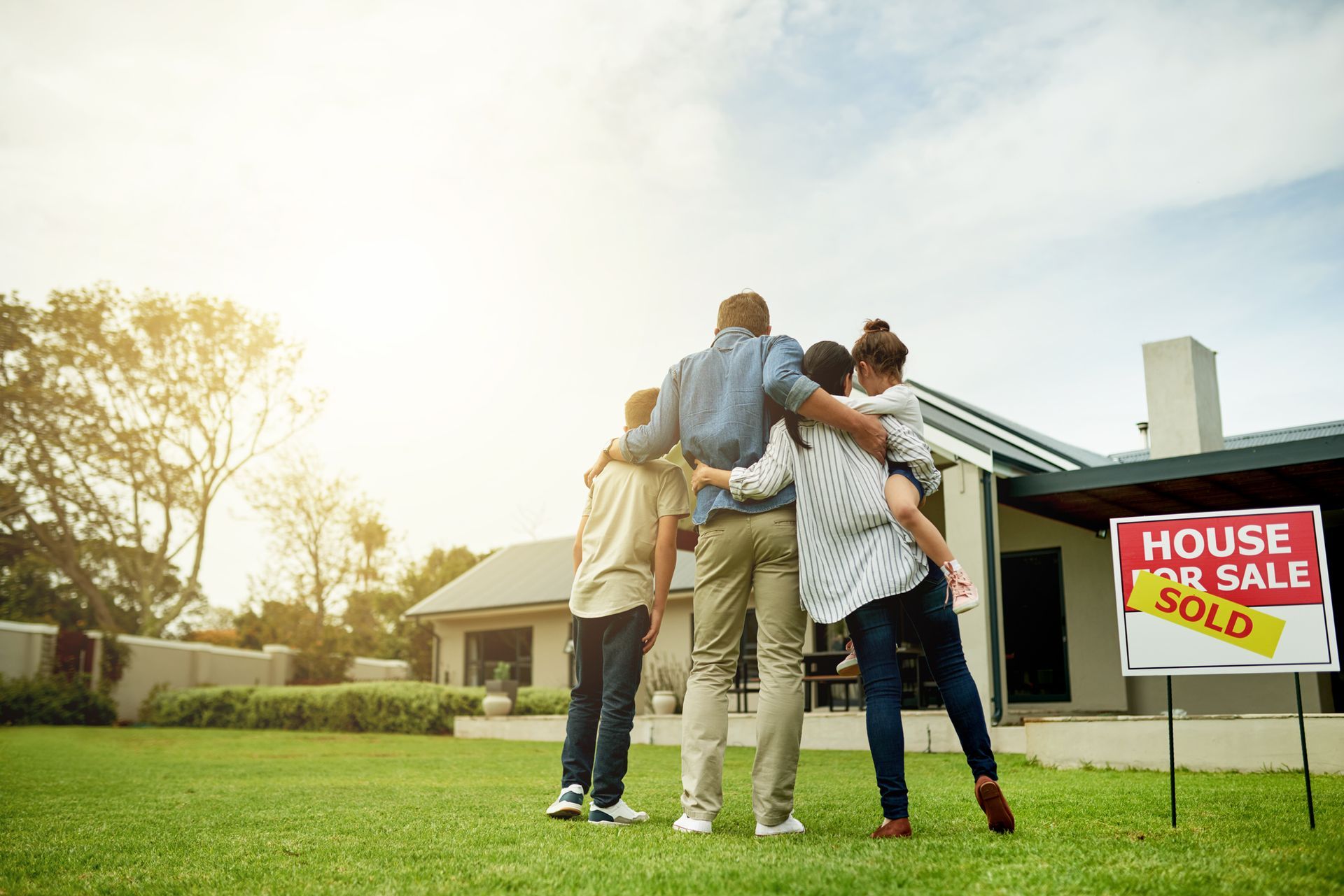 A family is hugging in front of a house for sale sign.