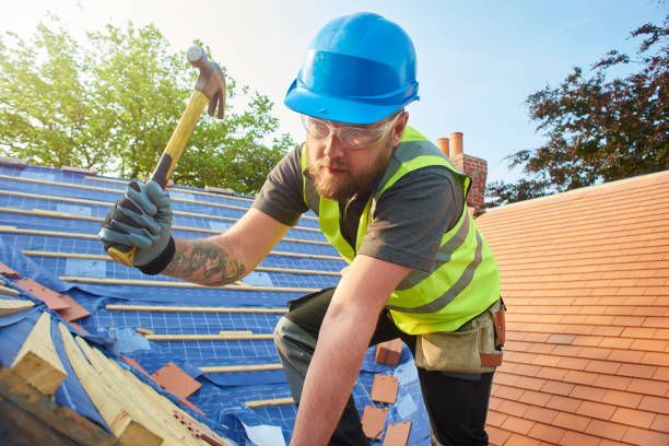 A roofer wearing a hard hat on a roof using a hammer during roof replacement on a building.