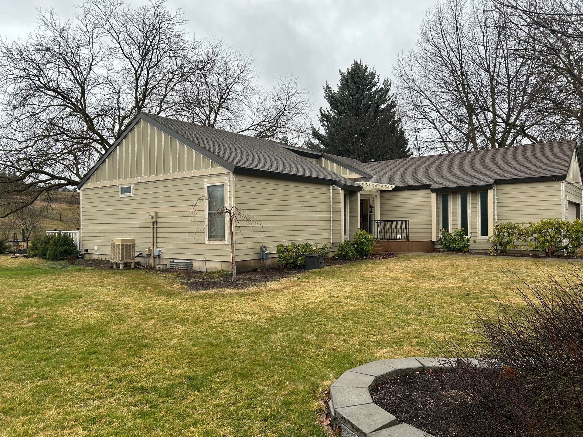 Beige house with a brown roof and brown trim sits on a grassy yard, bare trees in the background.