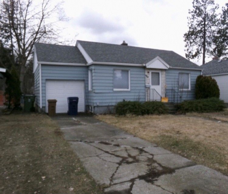 Light blue house with a gray roof and attached garage, cracked driveway, and small front yard.