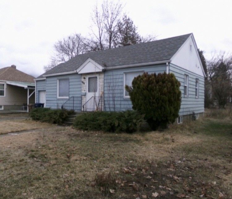 Blue-sided, single-story house with overgrown lawn and small shrub in front on a cloudy day.