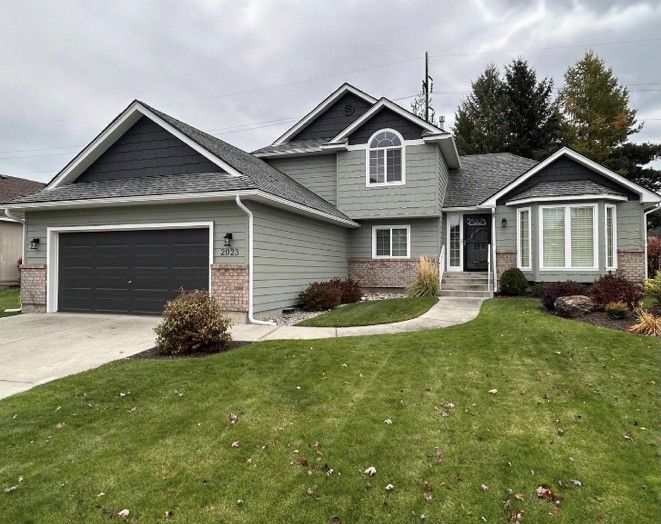 Green house with dark gray roof and garage door, brick accents, and a front lawn.