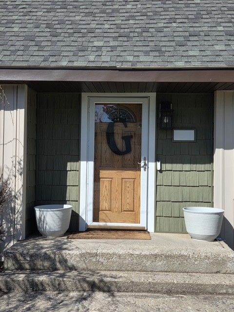 A wooden front door with a large letter G wreath, framed by olive green shingles, two white planters, and stone steps.