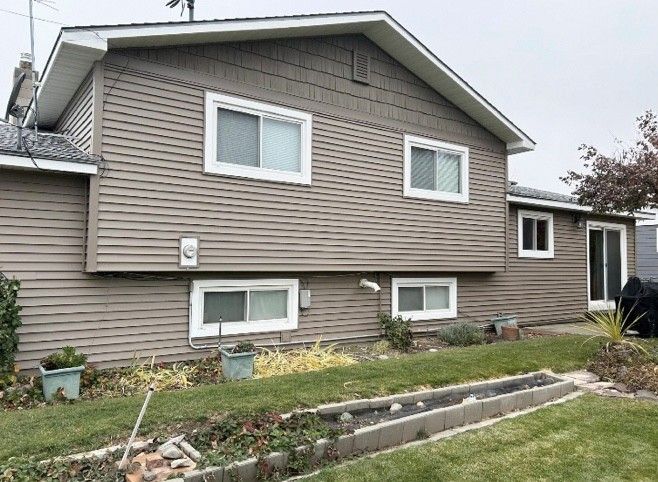 Brown house with white-framed windows, surrounded by grass and garden beds.