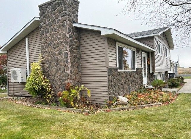 Tan house with stone chimney and foundation; green lawn and flower bed in front.