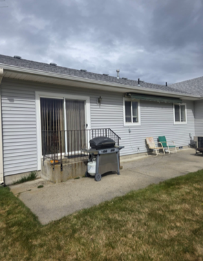 A back patio with gray siding, a sliding glass door, a grill, and two chairs under a cloudy sky.