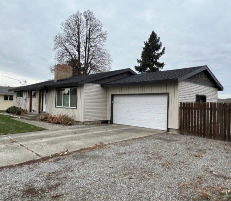 Tan ranch-style house with attached garage, gray roof, concrete driveway, and brown wooden fence. Overcast day.