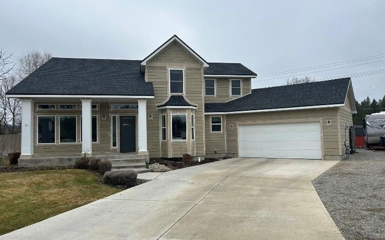 Tan two-story house with black roof, white trim, and a concrete driveway on a cloudy day.